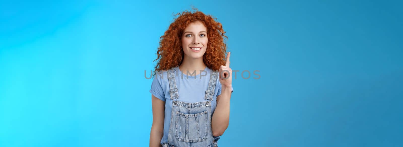 Cheerful cute energized redhead curly girl wear denim overalls book summer vacation trip offer good offer link pointing raised index finger up show uppwards advertisement smiling silly by Benzoix