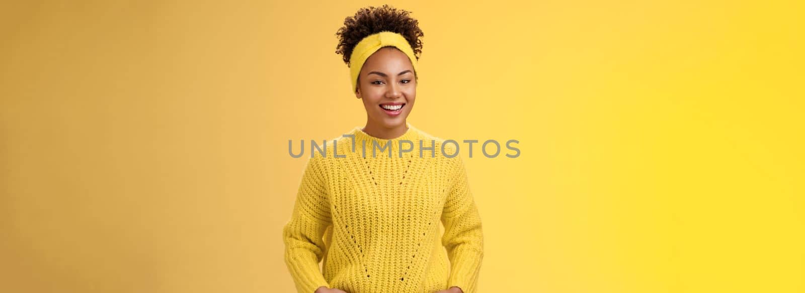 Friendly outgoing relaxed young african-american beautiful girl headband forehead in sweater hold hands pockets smiling broadly have fun talking interesting communication, standing yellow wall.