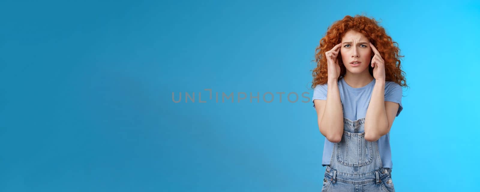 Confused panicking young distressed redhead curly girlfriend frowning touch temples cannot focus look perplexed worried not remember trying pull emotions together standing blue background.