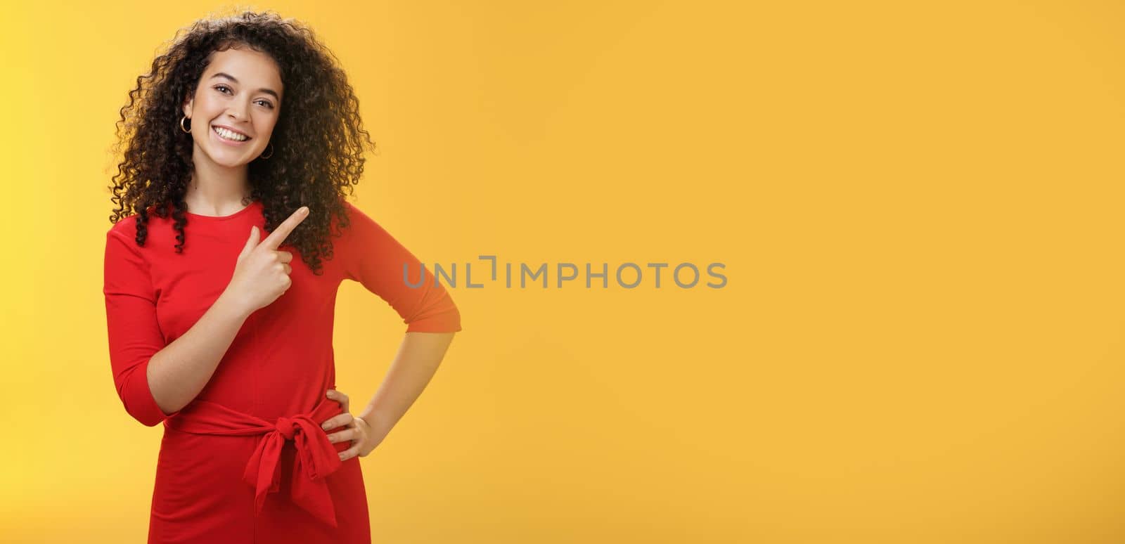 Girl suggesting try out new product. Friendly and pleasant happy young european brunette with curly hair tilting head cute, smiling broadly, pointing at upper right corner to direct at advertisement by Benzoix