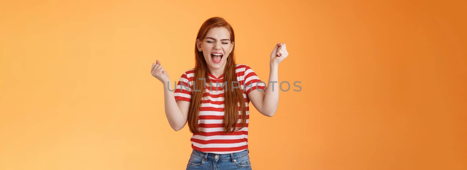 Luck cute redhead girl, make fist pump cheerfully yell yes, close eyes, winning lottery, feeling amazed, celebrate success and victory, stand orange background, triumphing, rejoicing good news by Benzoix