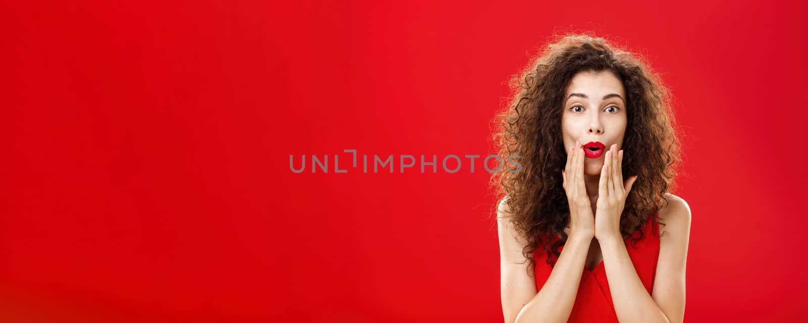 Charmed and intrigued curious good-looking rich woman in elegant dress with make-up and curly hairstyle folding lips in wow sign holding hands on face and smiling from excitement over red background by Benzoix