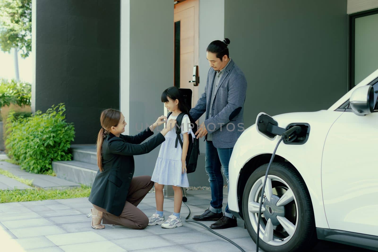 Progressive young parents and daughter with electric vehicle and home charging station. Green and clean energy from electric vehicles for healthy environment. Eco power from renewable source at home.