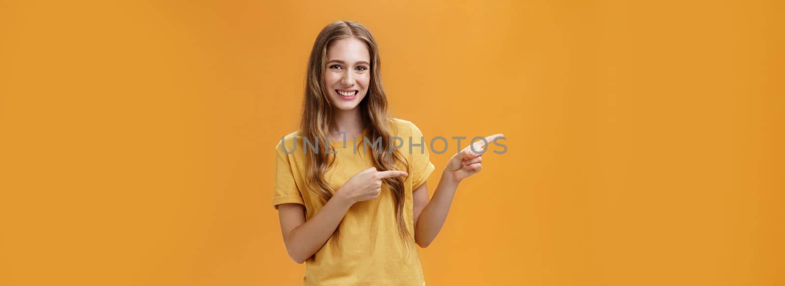 Girl shows cool place to visit. Portrait of charming carefree pretty young lady with cute natural wavy hair pointing left with index fingers and smiling broadly howing perfect place for advertisement.