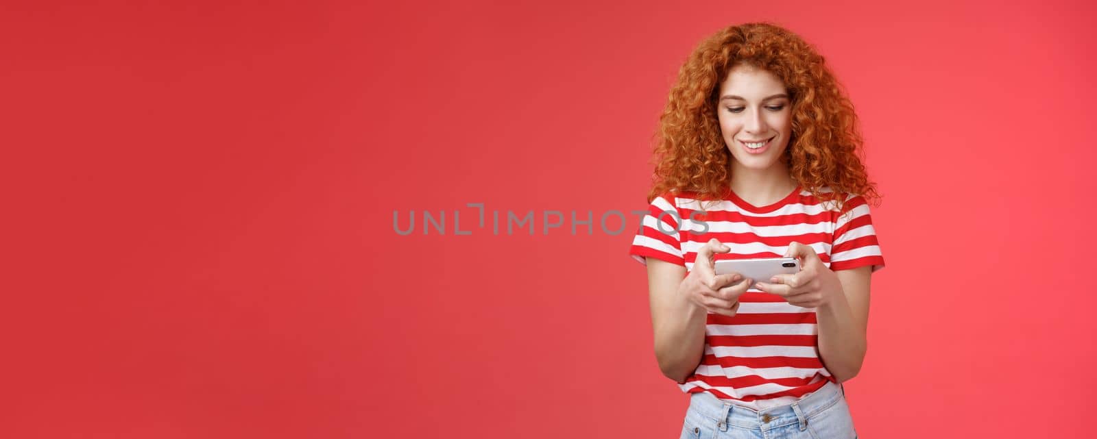 Redhead girl fool around waiting queue dentist playing awesome smartphone game hold phone horizontal tap cellphone screen look telephone display smiling delighted entertained standing red background by Benzoix