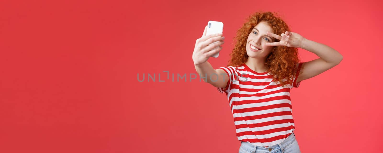 Hey followers how your summer holidays. Cheerful self-assured stylish fashionable redhead curly girl strike pose show peace victory gesture look smartphone screen taking selfie red background by Benzoix