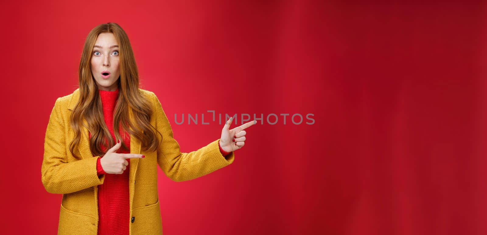 Speechless questioned and confused surprised redhead girl in yellow autumn coat pointing right open mouth curiously and staring at camera puzzled and clueless waiting explanation over red wall by Benzoix