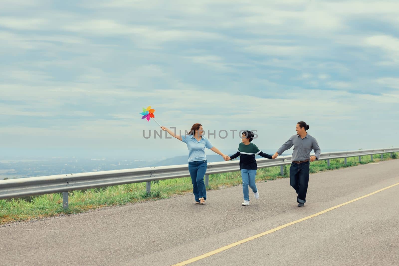 Progressive happy family vacation and carefree day concept. Young parents mother father and son run along and flying kite together road with enjoy natural scenic on scenery and clear sky background.