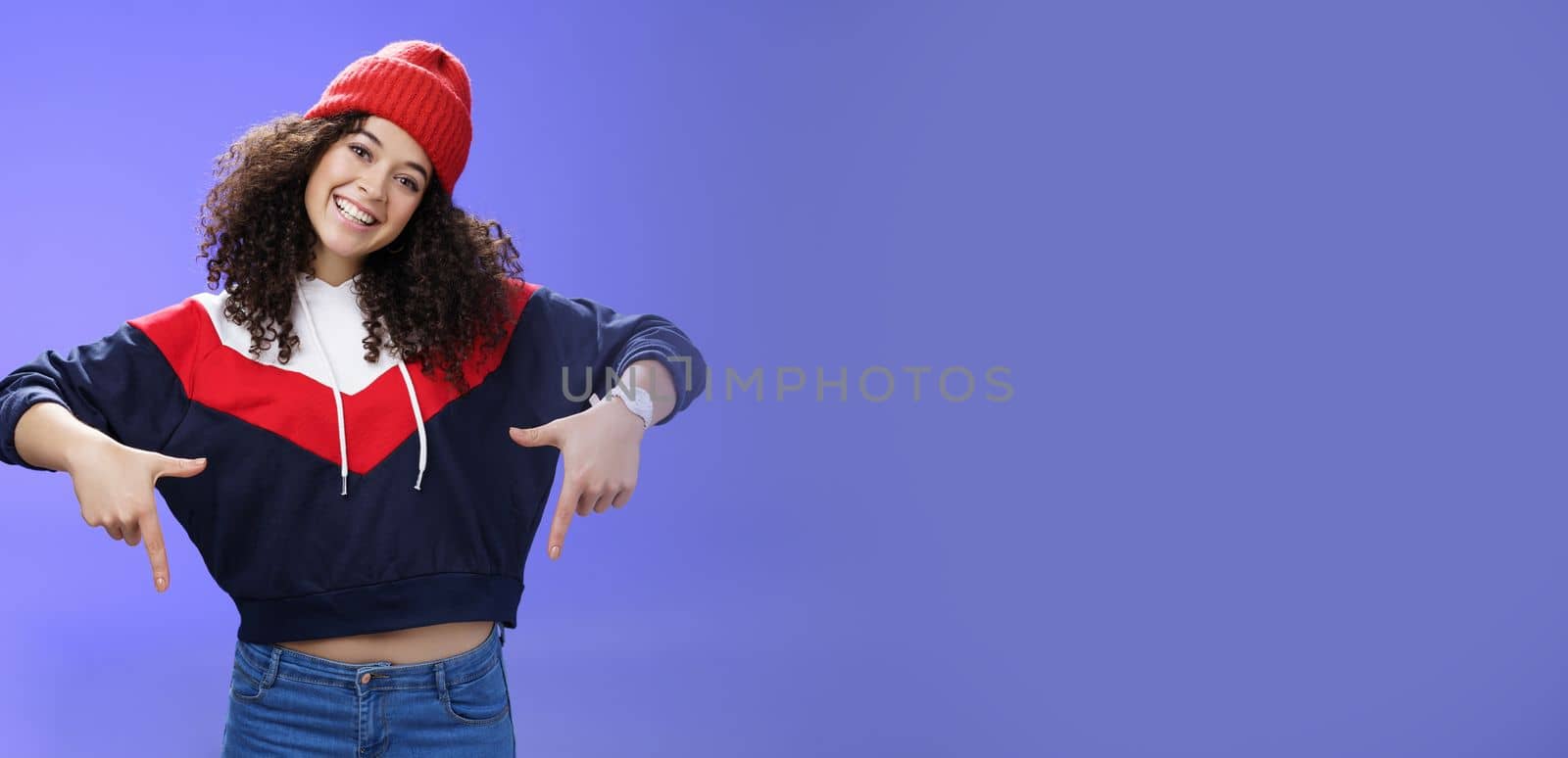 Waist-up shot of cool and stylish young woman with curly hair smiling flirty and joyful pointing down showing promotion as tilting head happily and posing over blue background in outdoor clothes by Benzoix