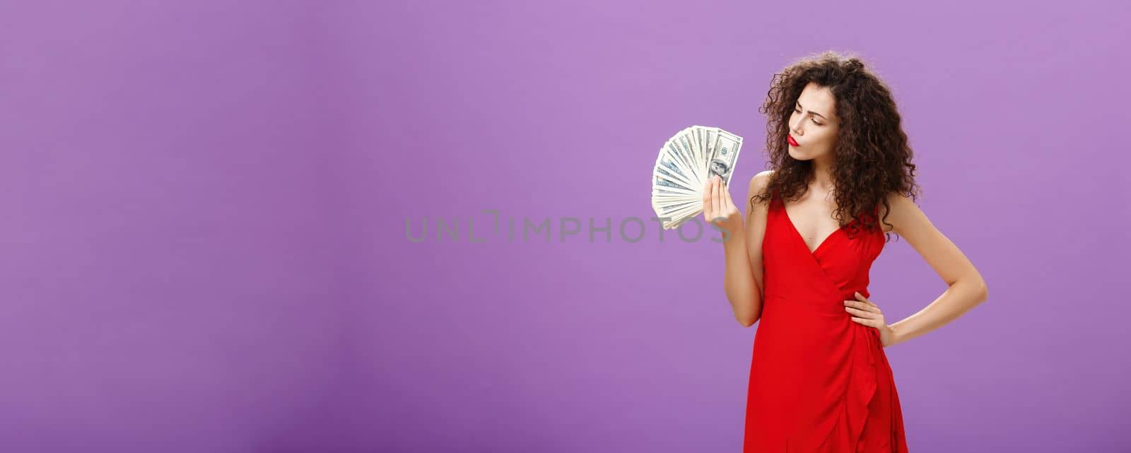 Woman deciding how spend lots of cash holding and looking at money. folding lips making decision or thinking how hard being rich standing elegant over purple background in red dress and curly hairstyle.