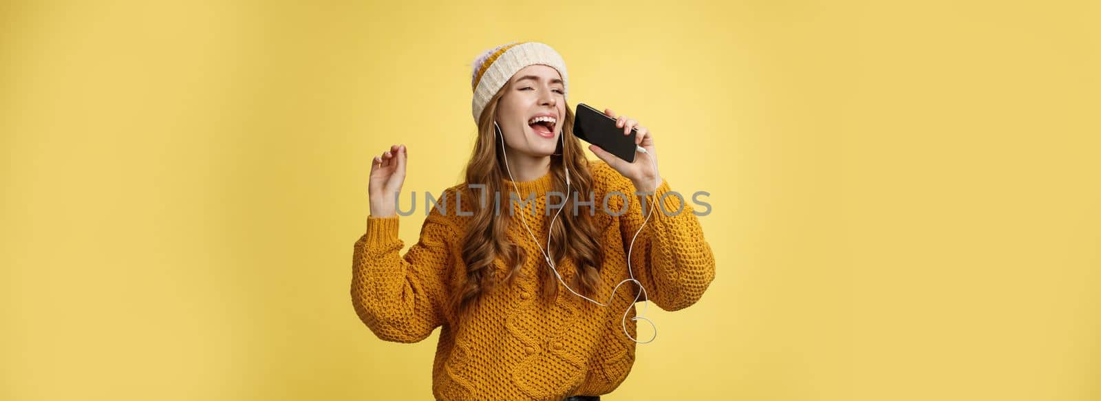 Carefree charming girl likes karaoke having fun listen favorite songs wearing wired earphones hold smartphone microphone singing out loud dancing enjoying spend time alone by Benzoix