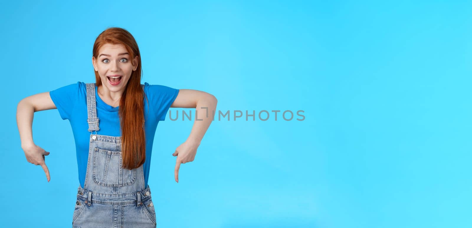 Amused wondered excited cute 20s european woman, pointing down, bottom copy space, look amazed admiration camera, describe amazing product, recommend site, stand blue background. Copy space