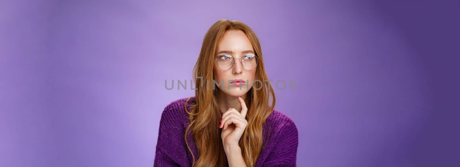 Girlfriend solving puzzle like detective making assumptions focusing on problem squinting looking left determined and thoughtful wearing glasses touching jaw while thinking over purple background.