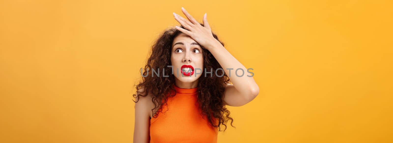 Shit I totally forgot about appointment. Nervous panicking displeased young european female with curly hair punching forehead with palm popping eyes anxiously at upper right corner making mistake.