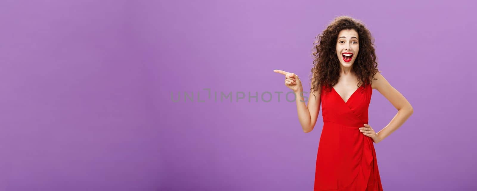 Portrait of entertained and amused joyful european woman. with curly hairstyle in red evening dress laughing from amusement and joy pointing left being entertained posing over purple background.