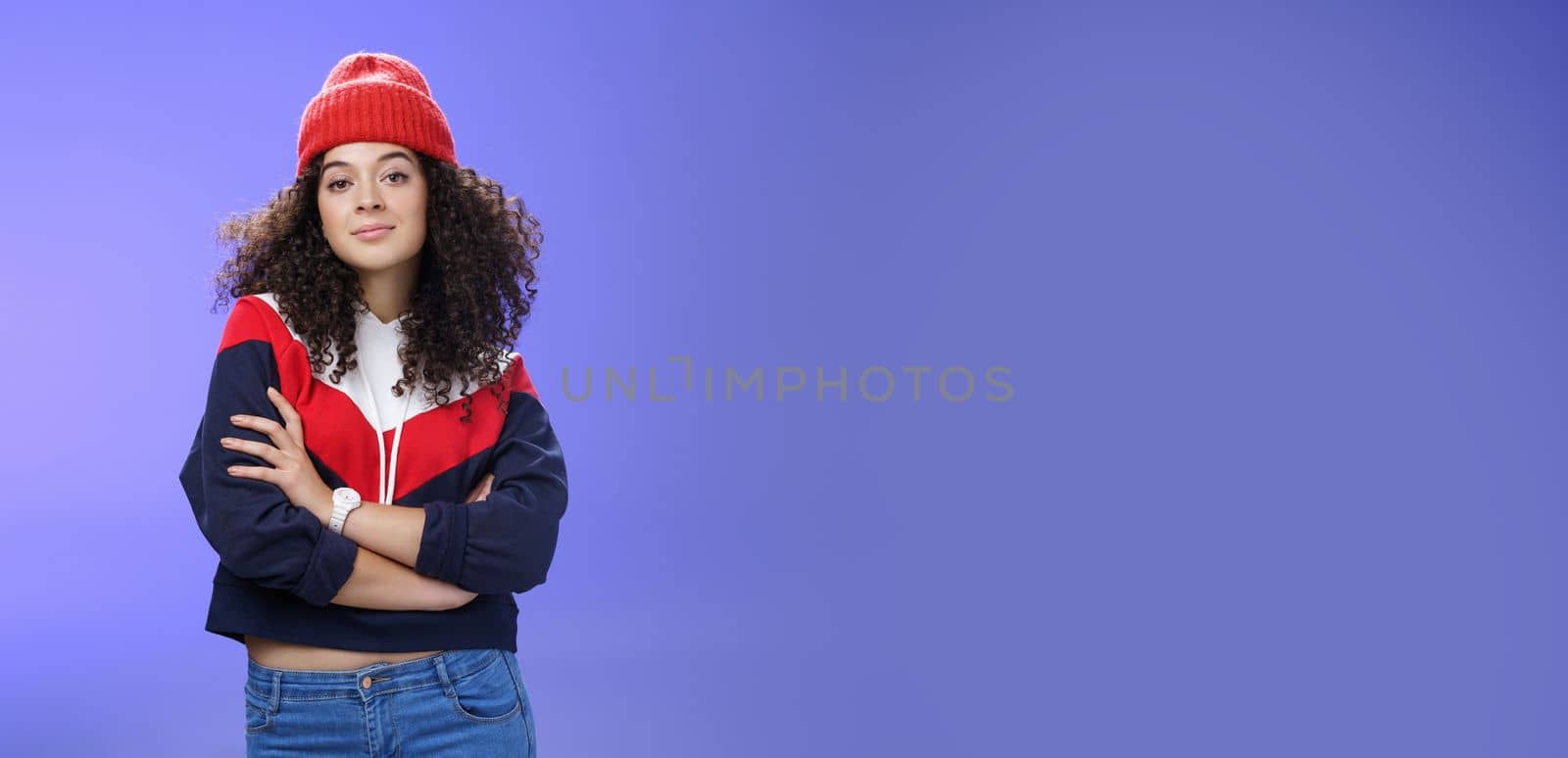 Girl will show winter who boss. Confident and stylish self-assured attractive woman with curly hair in cute red beanie and warm sweatshirt holding hands crossed over chest and looking daring at camera by Benzoix