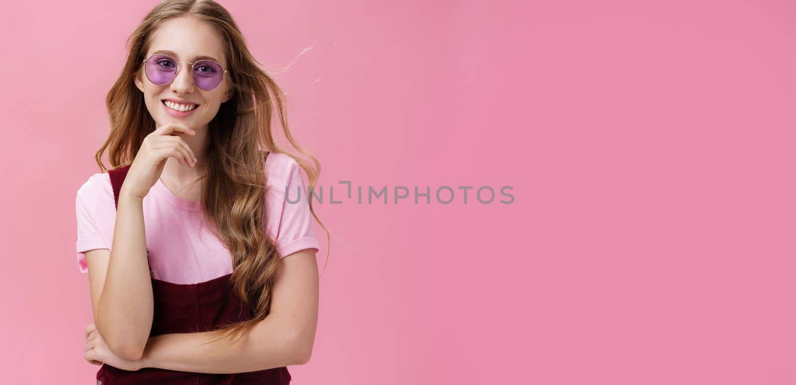 Beauty, style and fashion concept. Portrait of charming confident european female with natural wavy hair in sunglasses and overalls touching chin amused and smiling broadly against pink wall.