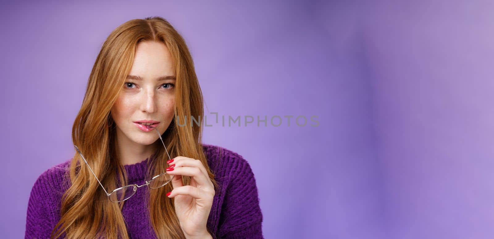 Close-up shot of smart and creative female redhead genius biting frame of glasses and squinting curious at camera as thinking having idea, making up plan against purple background.