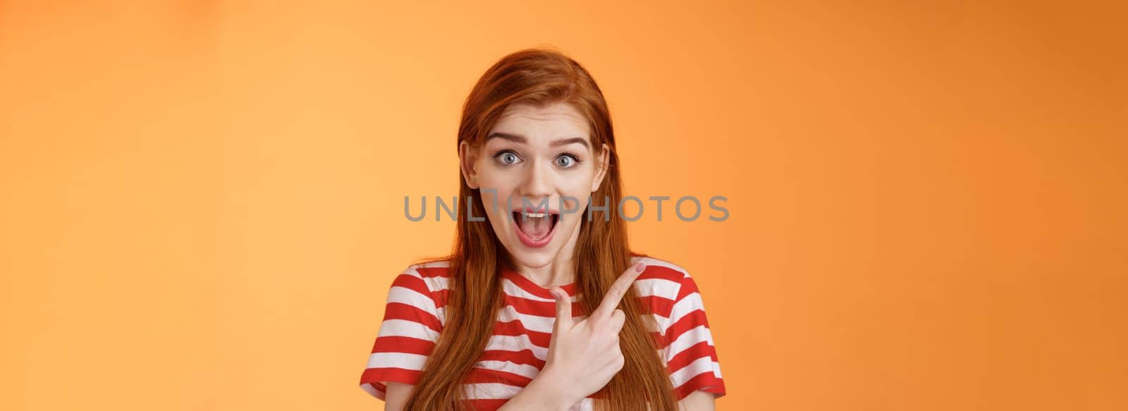 Excited female redhead hear impressive sale offer pointing upper left corner, drop jaw astonished stare camera full disbelief surprise, discuss interesting promotion, stand orange background.