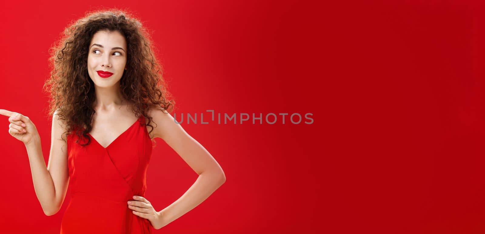 Stylish and elegant curious charming woman. with curly hairstyle in red lipstick and trendy dress holding hand on hip pointing and looking left delighted and interested over studio background.