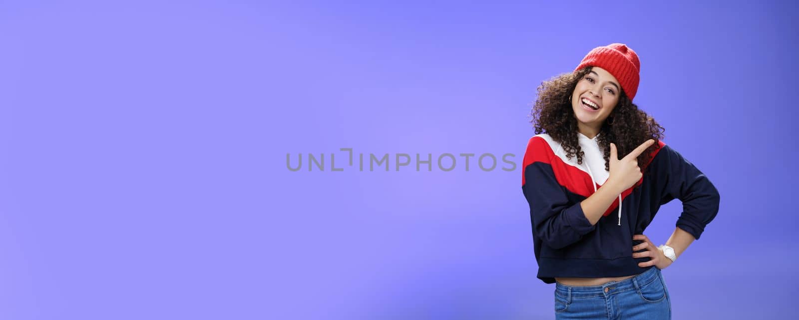 Studio shot of enthusiastic charming curly-haired woman inviting to play in snow with her pointing at upper right corner tilting head joyfully and smiling, holding hand on waist over blue wall by Benzoix