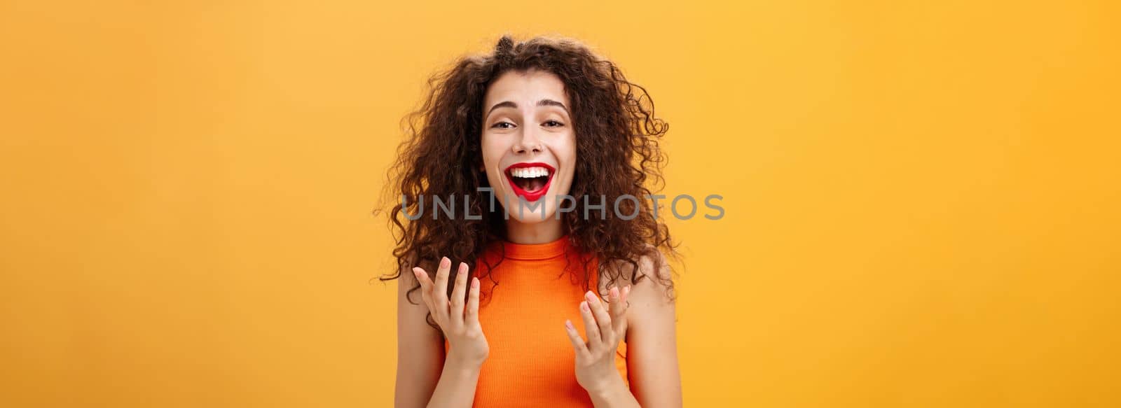 Surprised thrilled and joyful charming european female. with curly hairstyle raising palms in grateful and delighted gesture smiling broadly hearing unbelievable perfect news over orange wall. Copy space