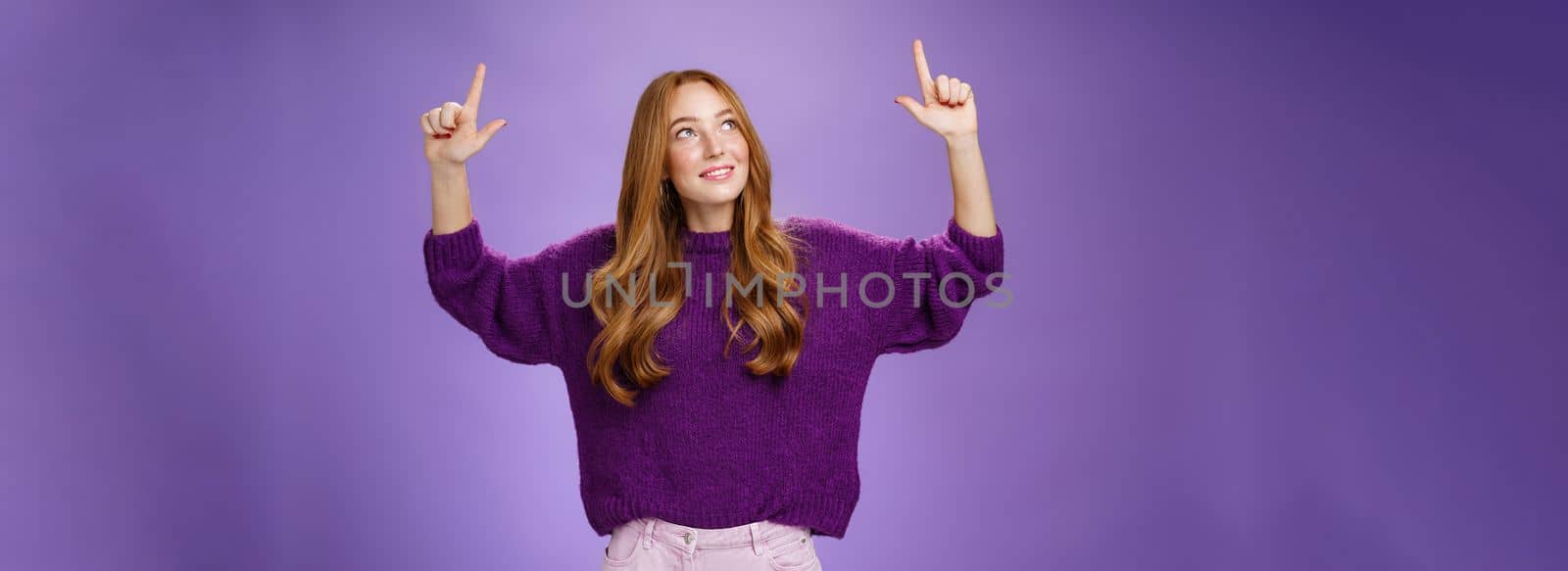 Lifestyle. Dreamy girl with red hair and freckles in warm cozy purple sweater raising hands looking and pointing up with intrigued and happy charmed expression smiling as liking curious product over violet wall.