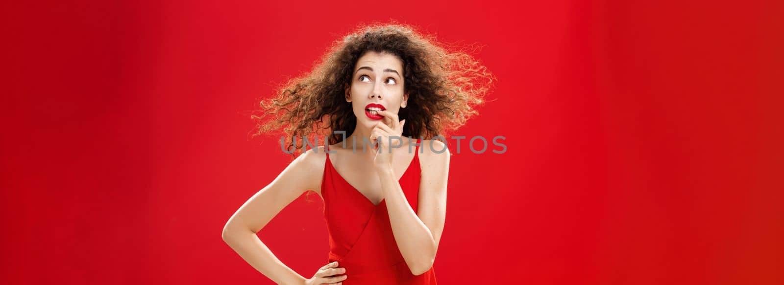 Waist-up shot of concerned and worried insecure cute female with curly hairstyle in red evening dress biting finger looking troubled or questioned at upper right corner with hand on waist. Copy space
