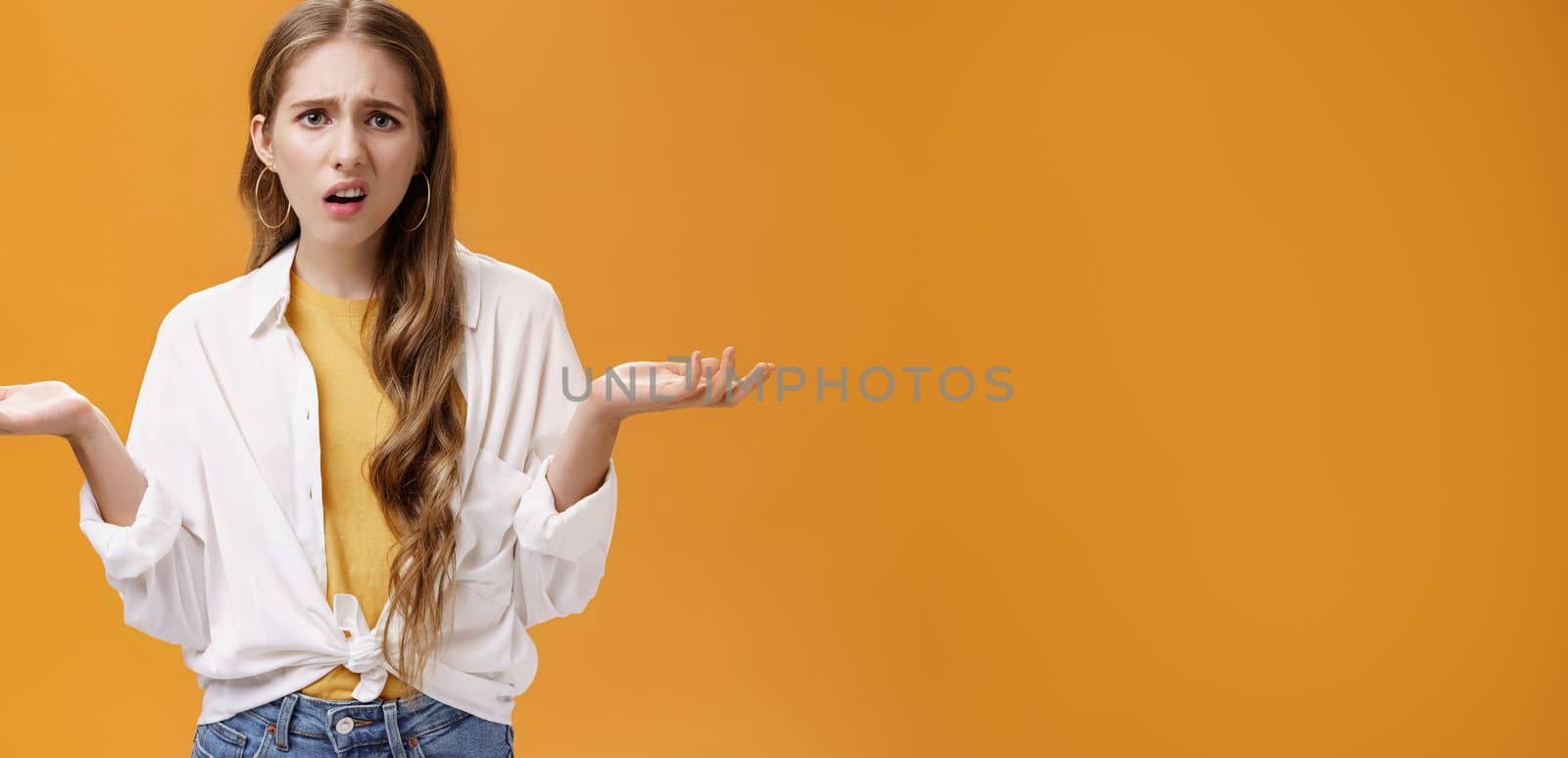 What, I do not understand. Portrait of arrogant questioned impolite girl being pissed with questiones shrugging with hands spread sideways making irritated grimace standing confused over orange wall.