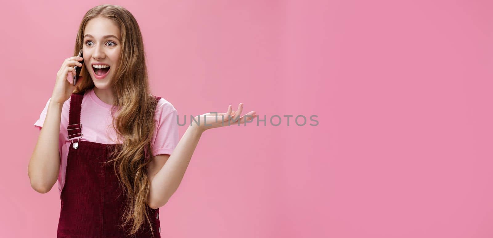 Portrait of popular outgoing and talkative young attractive girl in overalls waving raised hand during conversation talking on smartphone amused and excited gossiping with friend via cellphone by Benzoix