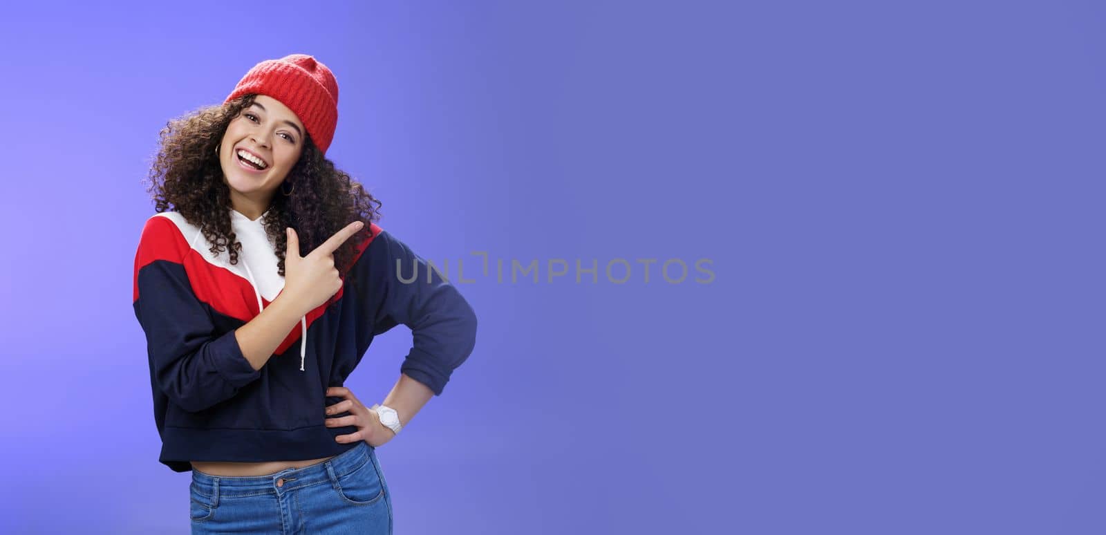 Studio shot of enthusiastic charming curly-haired woman inviting to play in snow with her pointing at upper right corner tilting head joyfully and smiling, holding hand on waist over blue wall. Emotions and weather concept
