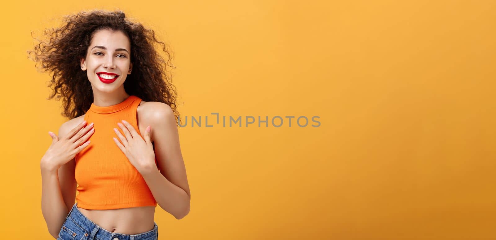 Waist-up shot of sensual and feminine tender female with curly hairstyle in cropped top holding palms on chest delighted and pleased smiling broadly posing flirty and sexy over orange wall. Copy space