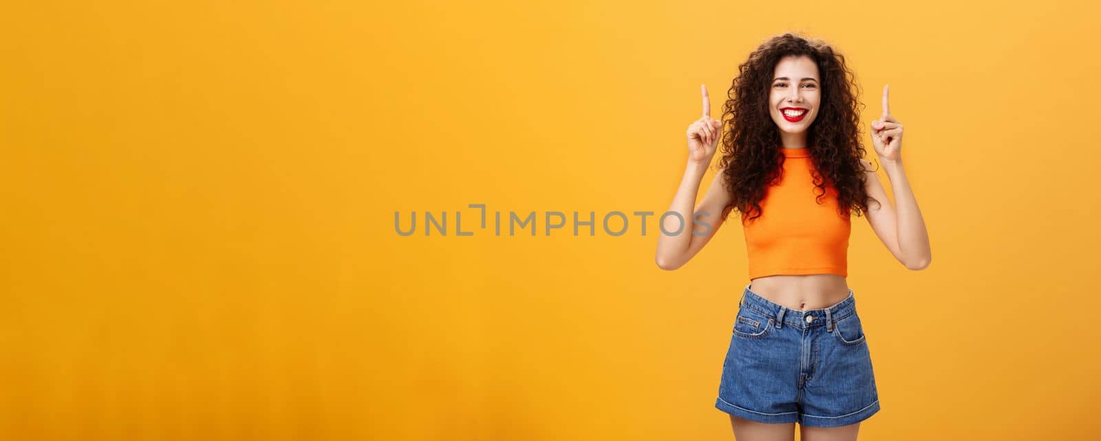 Indoor shot of friendly and cute happy relaxed female with red lipstick and curly hairstyle wearing cool cropped top and denim shorts smiling broadly pointing up posing over orange background. Copy space