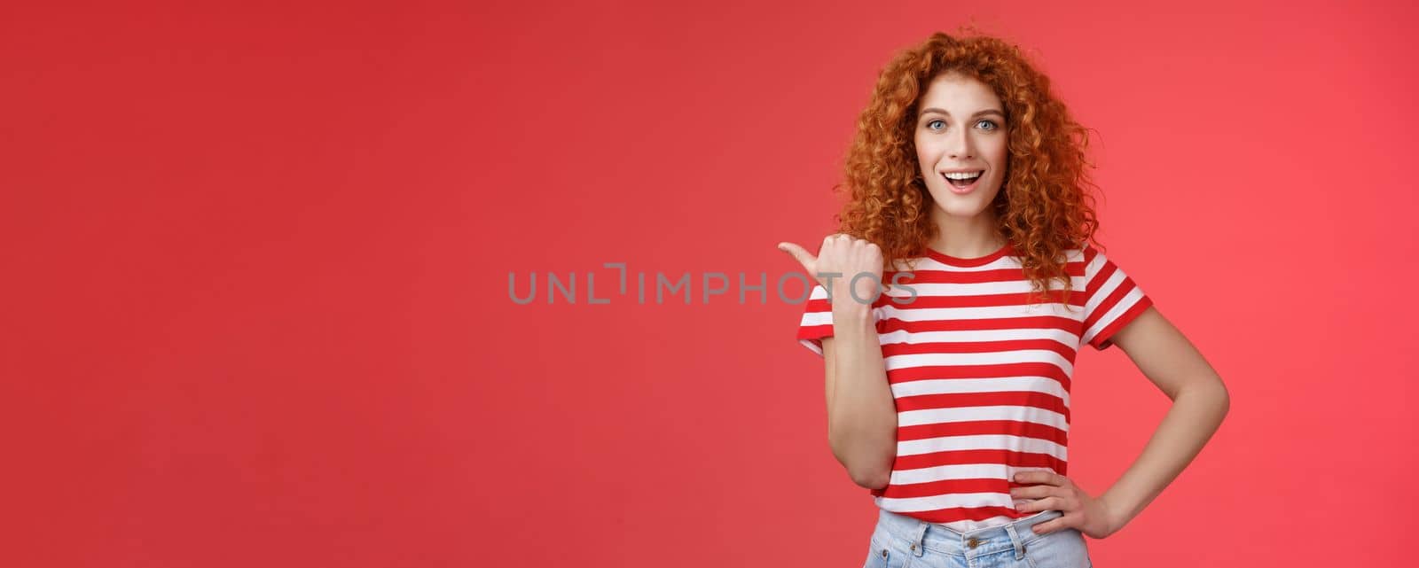 Lifestyle. Sassy good-looking emotive happy smiling redhead european female curly hairstyle pointing thumb left grin assertive cheeky hold hand waist directing promo advertising offer red background.