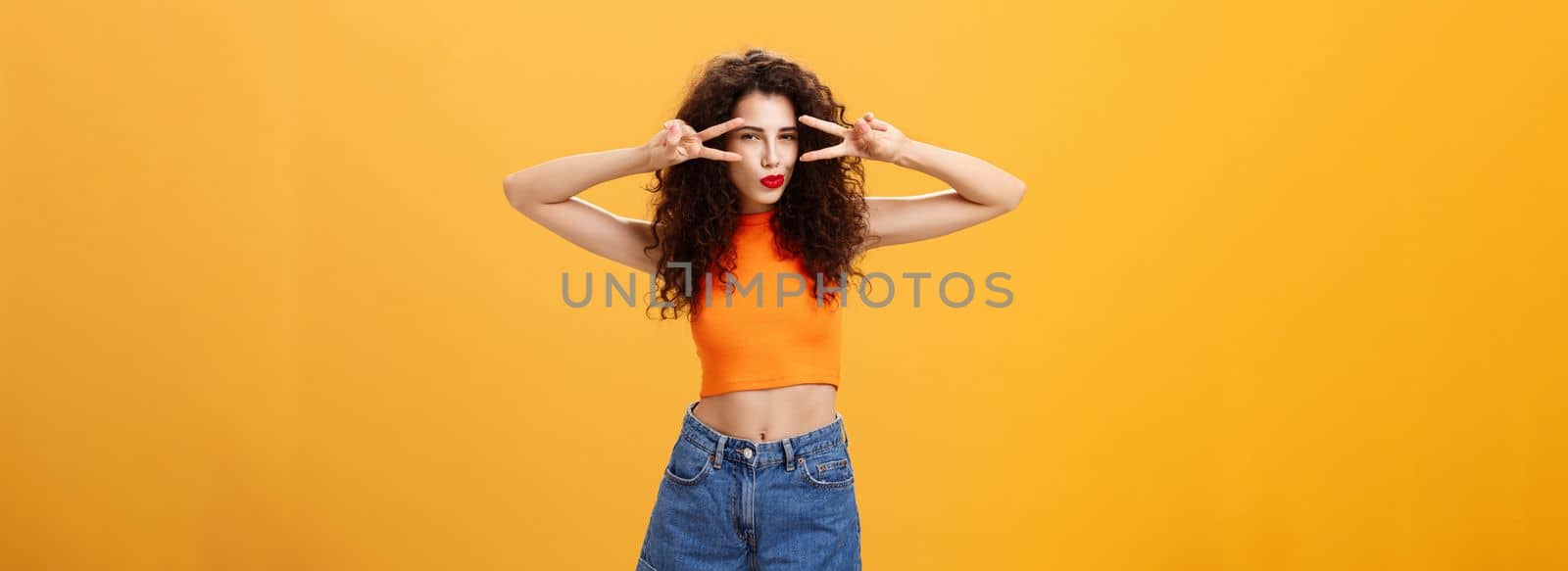 Portrait of good-looking daring and sexy stylish woman. with curly hairstyle in red lipstick and cropped top showing victory or peace gestures overs eyes squinting folding lips flirty over orange wall.