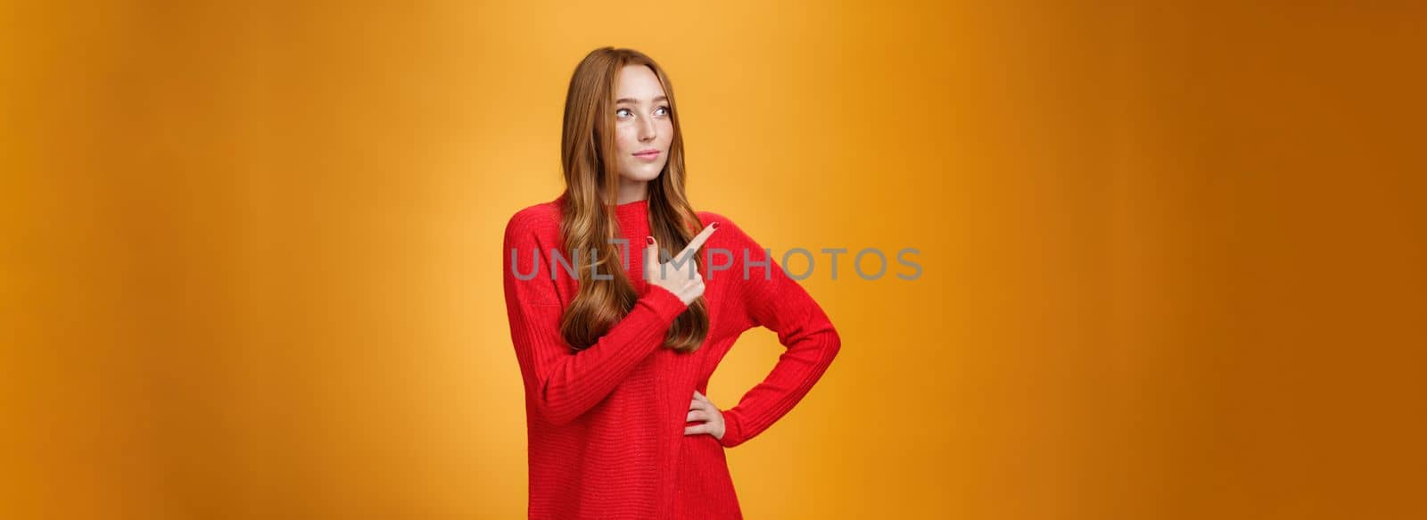 Portrait of glamour and stylish redhead female entrepreneur designing and leading new project pointing at upper left corner as gazing thoughtful and focused on job, managing business over orange wall.