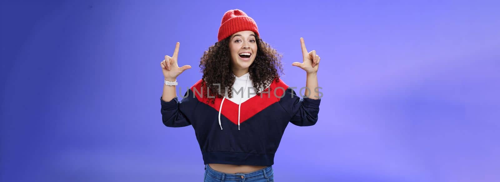 Lifestyle. Waist-up shot of cheerful energized young woman calling friend inviting hang out as weather awesome pointing up with raised hands smiling broadly posing in red winter hat over blue background.