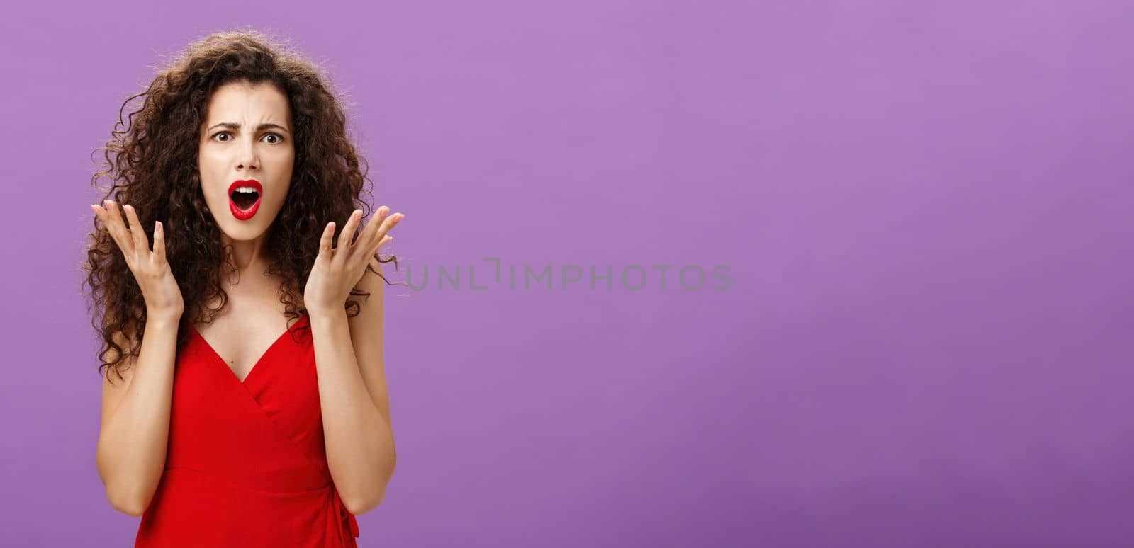 Rich arrogant and snobbish european woman. with curly hairstyle in red evening dress arguing with made frowning looking confused and displeased shaking palms in disappointment posing over purple wall.