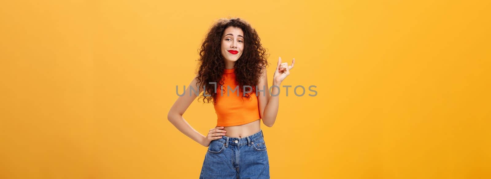 Girl needs bigger size to impress. Carefree picky stylish urban female with curly hairstyle red lipstick in cropped top shaping small or little thing with fingers smirking unimpressed over orange wall.