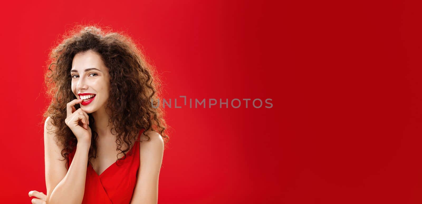 Waist-up shot of sexy and sensual adult girlfriend. with curly hair in elegant dress biting finger and smiling flirty seducing hot guy during formal event posing against red background romantically.