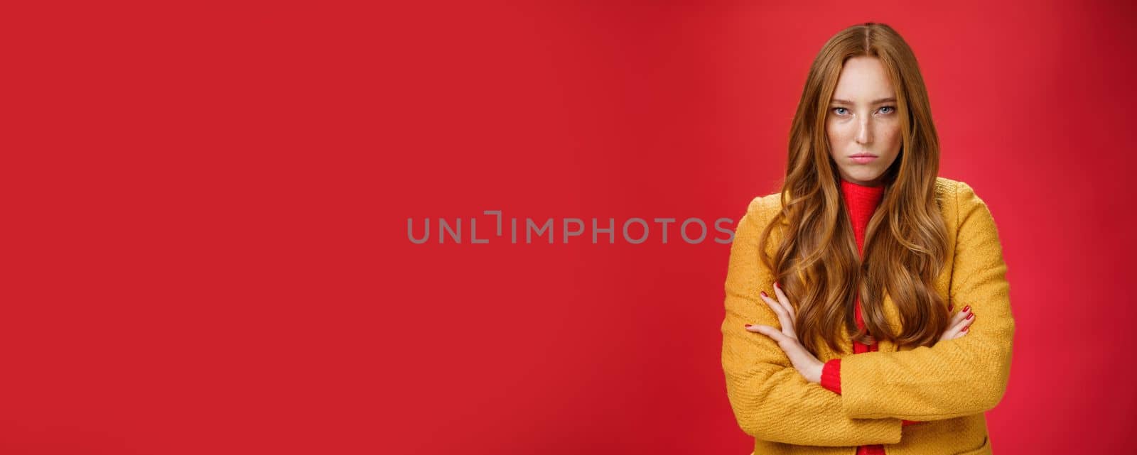 Offended and angry redhead girlfriend expressing dismay and hate as being waiting too long crossing hands over chest in defensive and aggressive pose, pouting looking with contempt over red background.