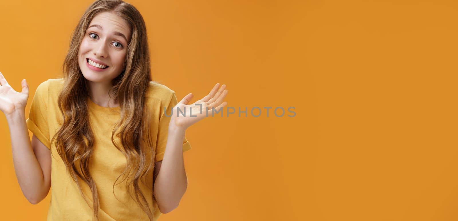 Girl stay out of business shrugging with hands raised and spread aside, silly sorry smile posing over orange background unaware and confused in casual t-shirt having no idea about topic.