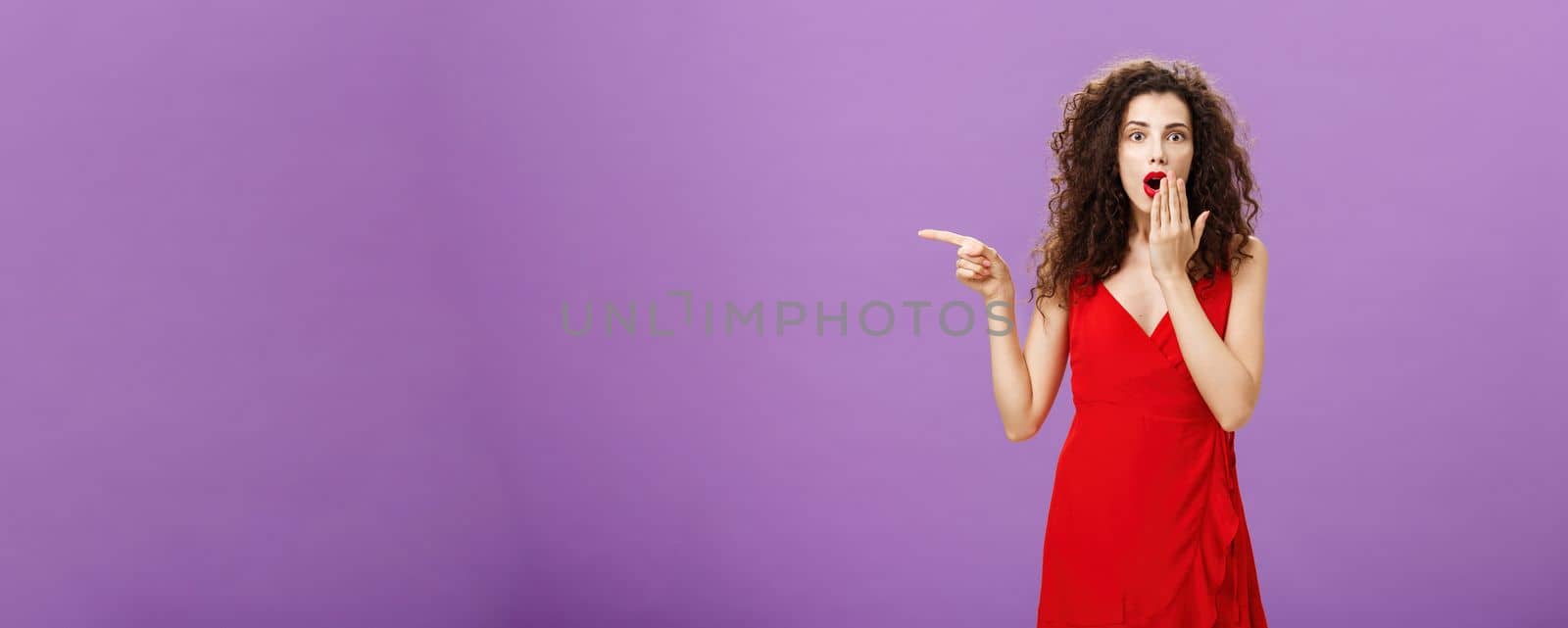 Portrait of surprised and amazed charming elegant lady. with evening make-up in red dress covering opened mouth from shock and amazement pointing left curious and questioned over purple background.