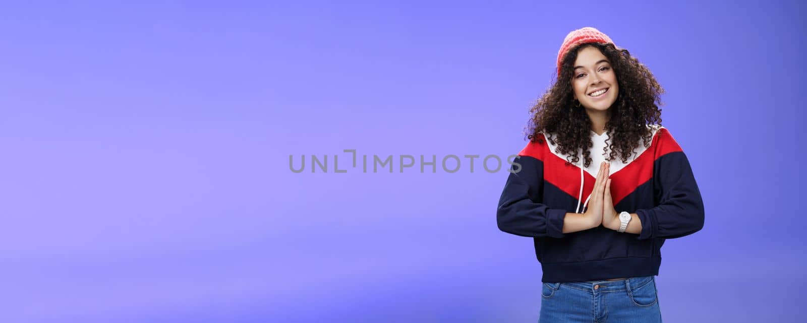 Portrait of charming curly-haired feminine girlfriend begging for favour as posing over blue background in winter outfit with hat, holding hands in pray smiling with angel look and friendly gaze by Benzoix