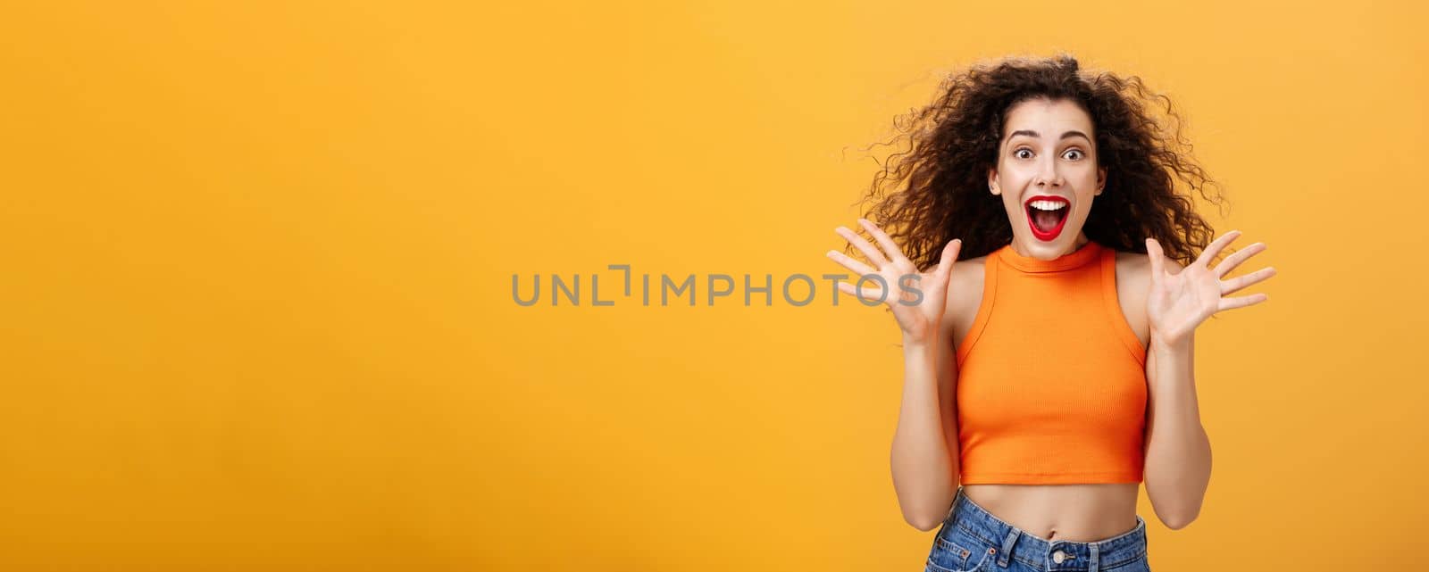 Excited impressed attractive stylish woman with nose ring, red lipstick and curly hairstyle smiling amazed with opened mouth gesturing with raised palms being astonished over orange background. Emotions concept