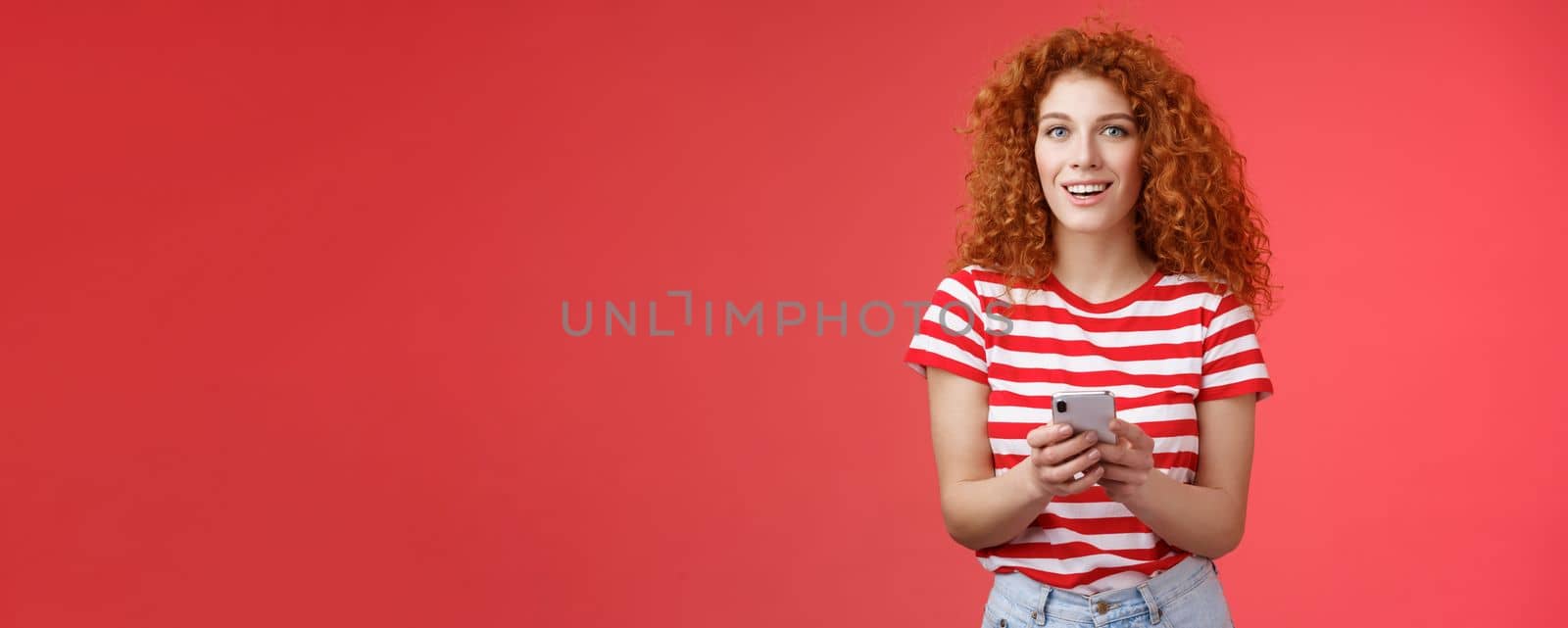 Fashionable carefree good-looking relaxed redhead girlfriend hold smartphone look camera excited pleased turn attention friend answering messages social media writing post red background by Benzoix