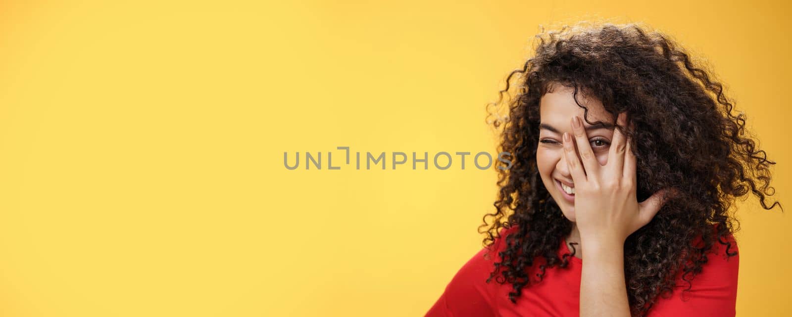 Close-up shot of sensual and playful attractive girlfriend with curly hair covering face with palm and peeking through fingers with happy tender smile anticipating surprise over yellow background by Benzoix