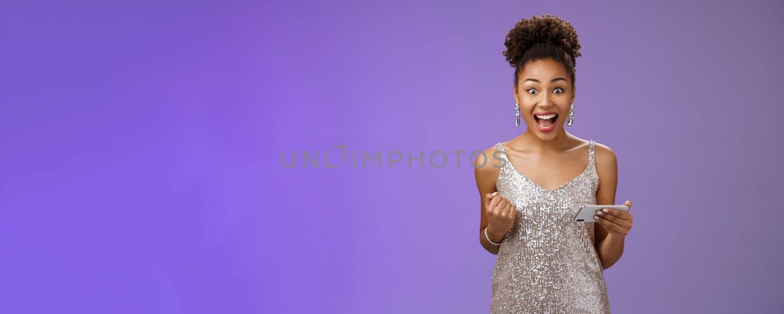 Excited charming young african-american woman lucky winning game playing smartphone standing pleased yelling happily clench fist triumphing joyfully celebrating great news received message by Benzoix