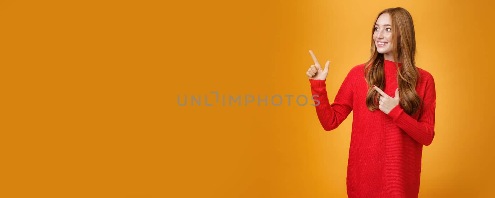 Interested gentle and tender young 20s ginger woman in red warm sweater pointing, looking at upper left corner with broad smile, enjoying nice happy scene over orange background. Advertisement concept