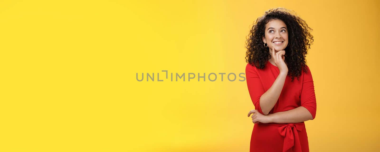 Girl having interesting idea, smiling as feeling confident plan might work. Charming happy woman with curly hair in red dress gazing at upper left corner thoughtful, thinking over yellow wall.